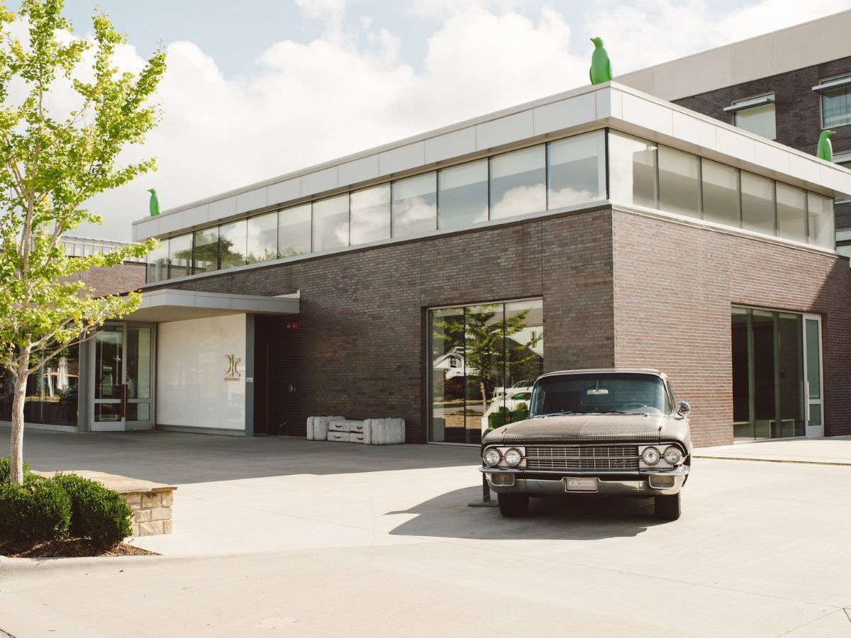 A vintage car is parked in front of a modern brick building with large windows and green sculptures on the roof. The sky is partly cloudy.
