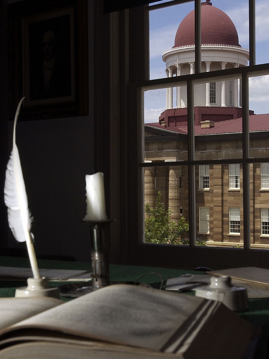 An open book and quill on a desk, with a candle beside them. The view through the window shows a building with a red dome roof.