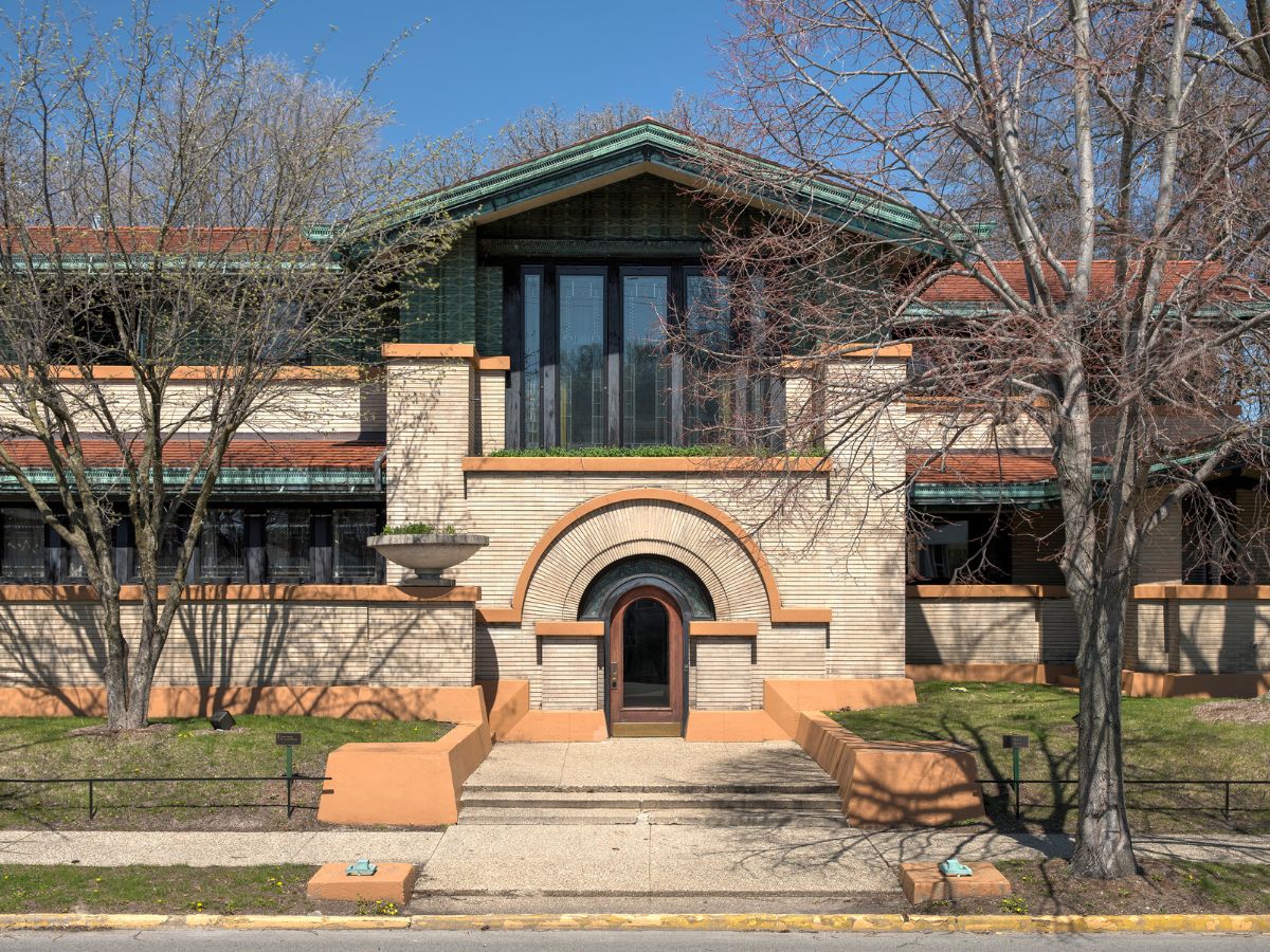 A two-story building featuring a light brick façade, large dark-framed windows, an arched entrance, and a red-tiled roof, with bare trees in the foreground and a paved pathway leading to the entrance.