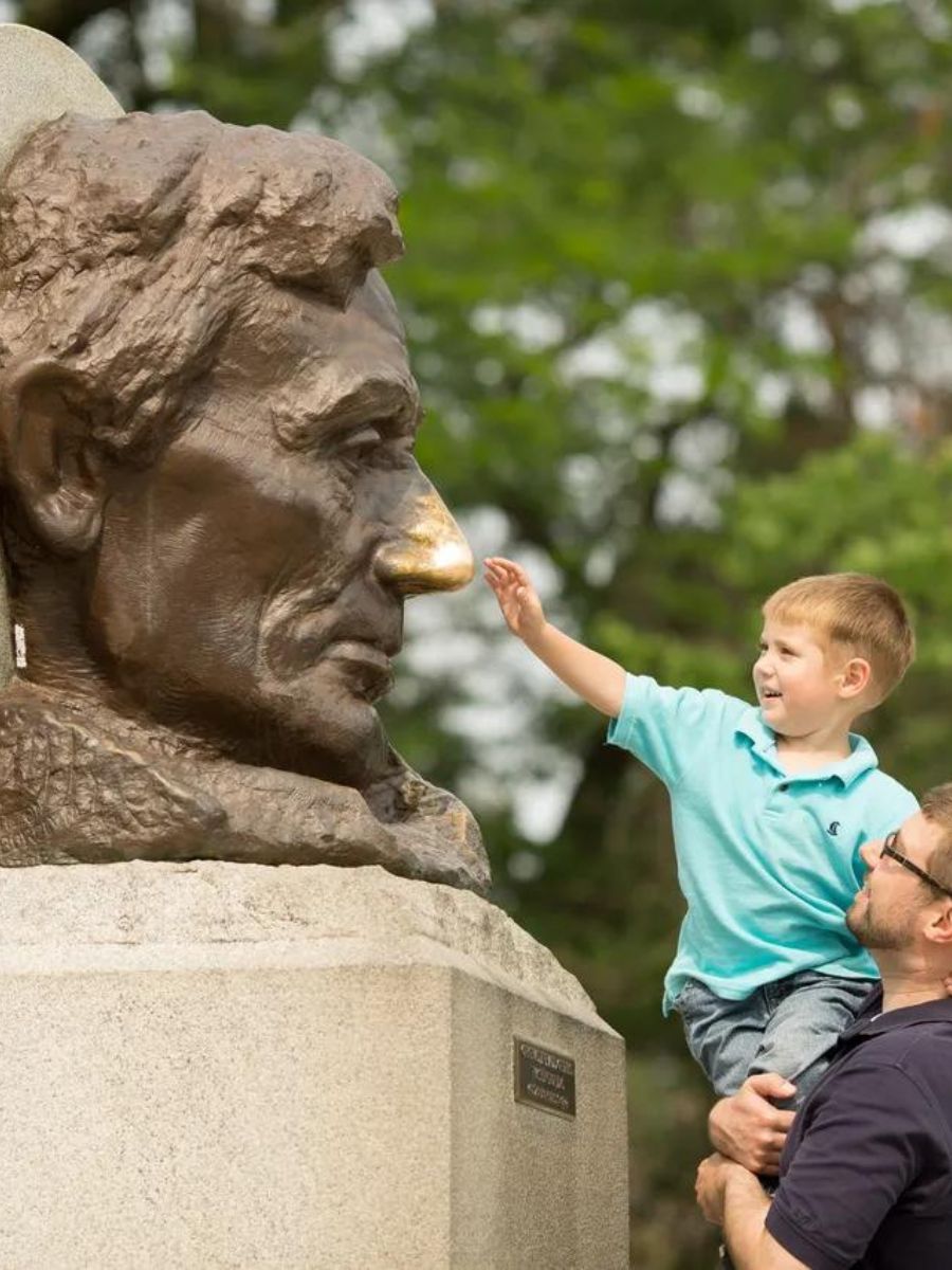 A child, held up by an adult, touches the nose of a large bronze statue's face outdoors.