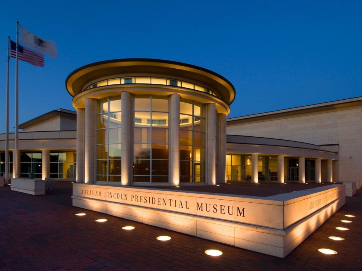 Exterior view of the Abraham Lincoln Presidential Museum at twilight, featuring a modern building with a cylindrical glass entrance and columns, with lit signage and two flags flying in front.