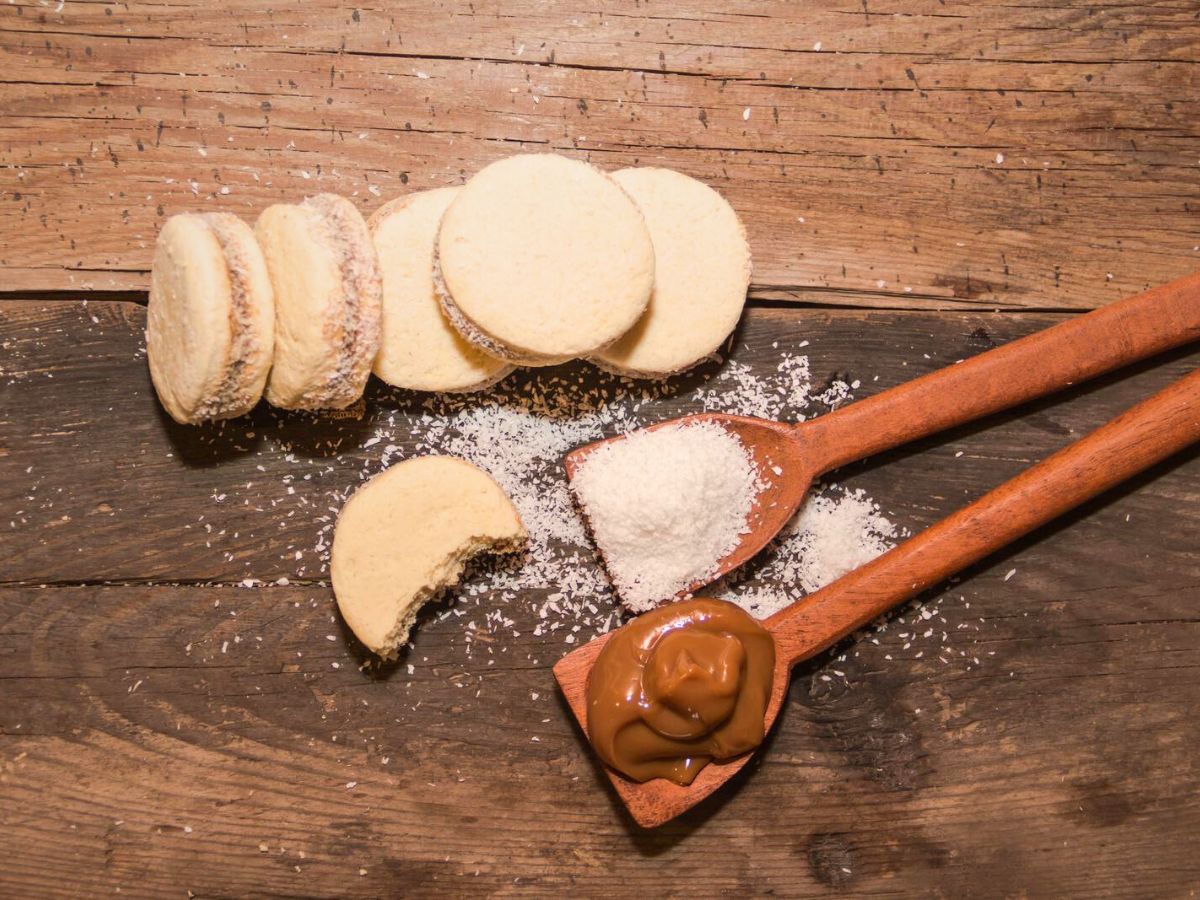 A few cookie sandwiches with one partially eaten, surrounded by shredded coconut and wooden spoons containing shredded coconut and caramel spread, all set on a rustic wooden surface.