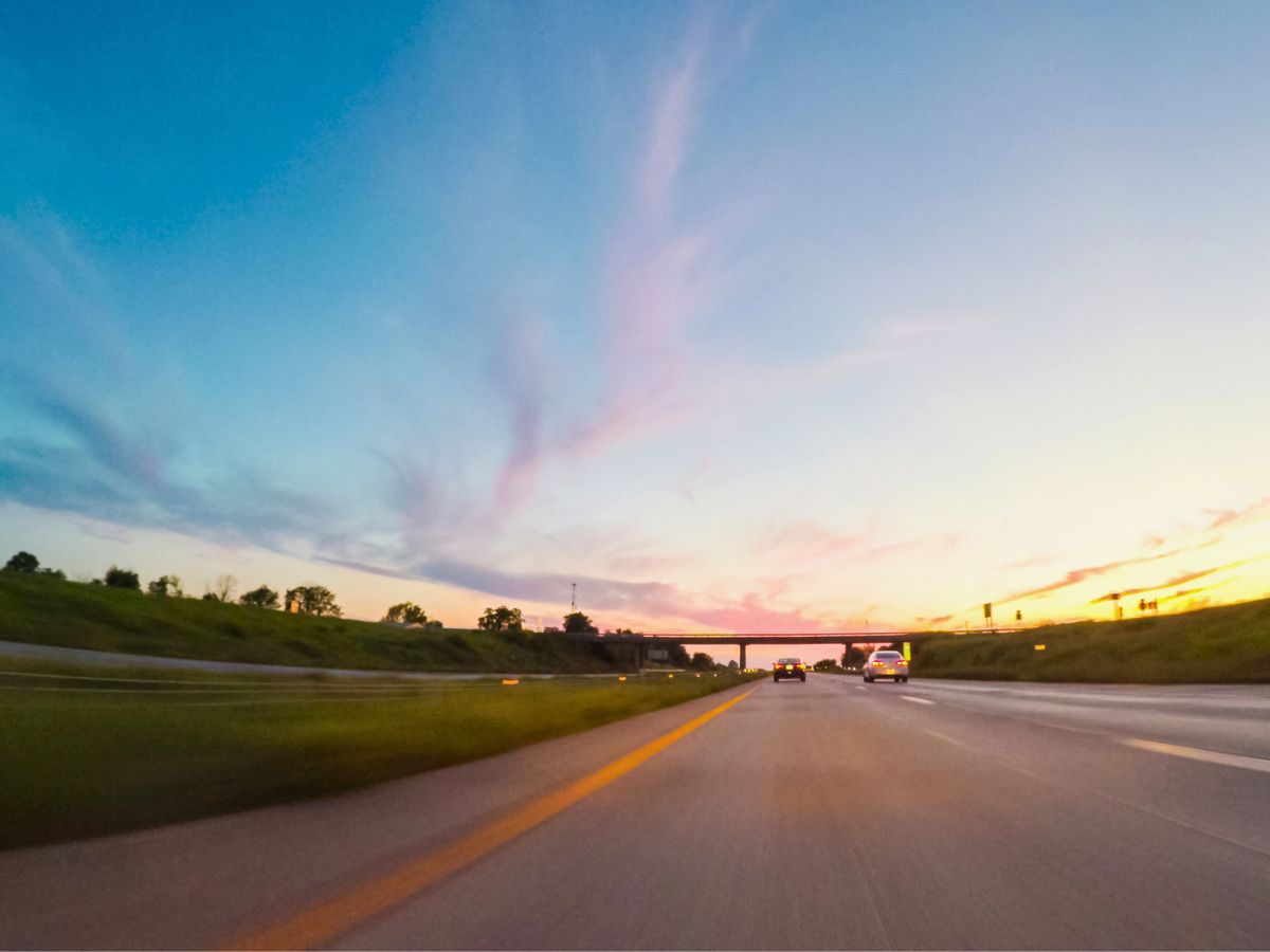 A view of a highway at sunset, with cars driving and a bridge in the distance, under a sky with blue and pink clouds.