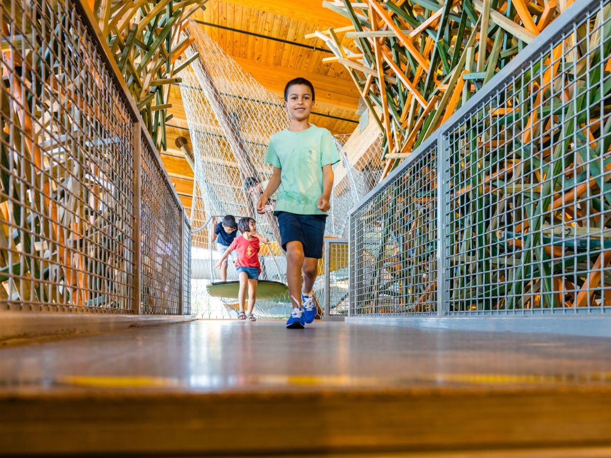 Two children walk across an indoor play structure with metal netting and wooden elements. The boy in front is in a green shirt, and the girl behind is wearing a red shirt.