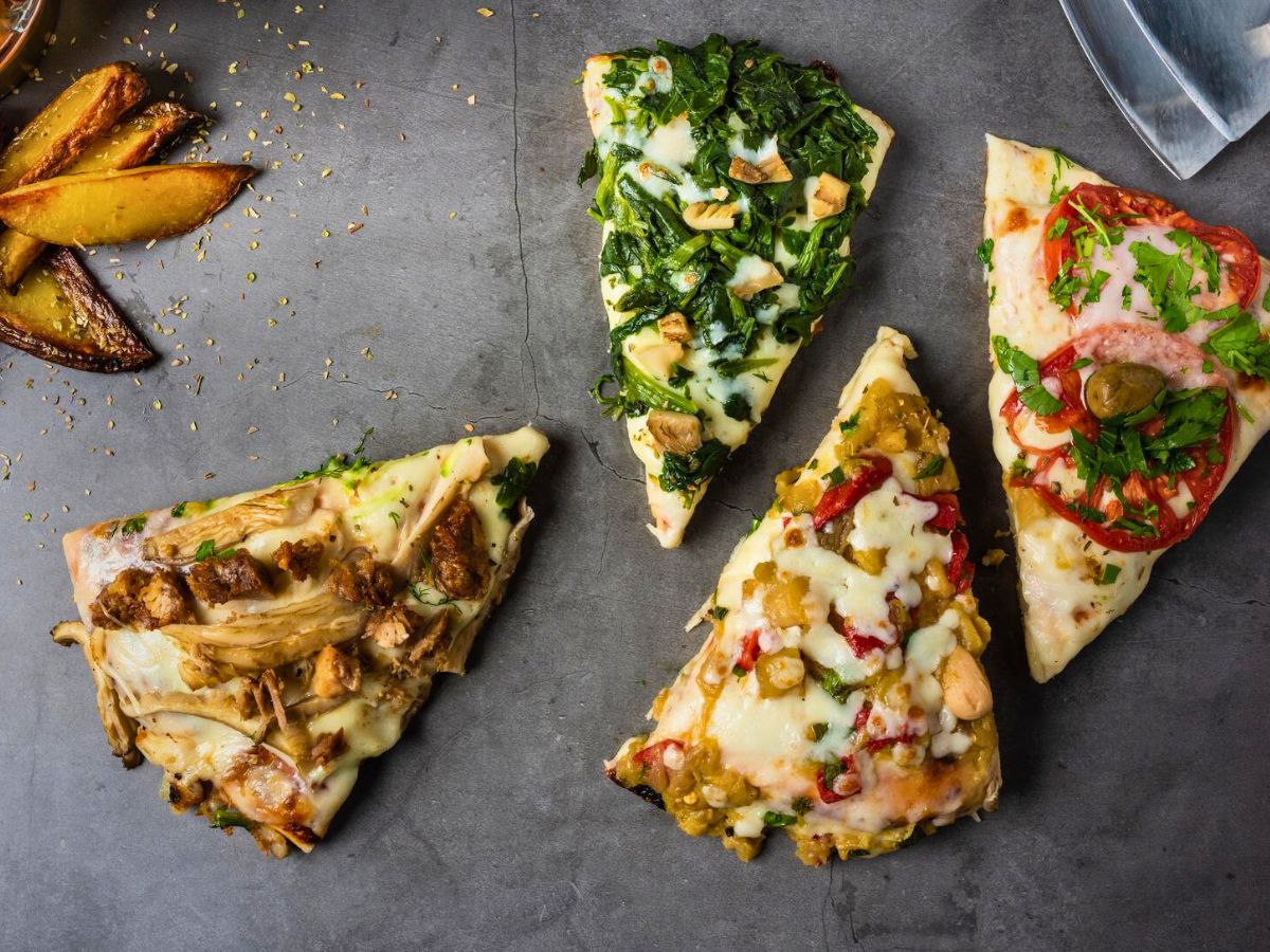 Four different slices of pizza with various toppings, arranged on a gray surface. There are three potato wedges in the upper left corner.