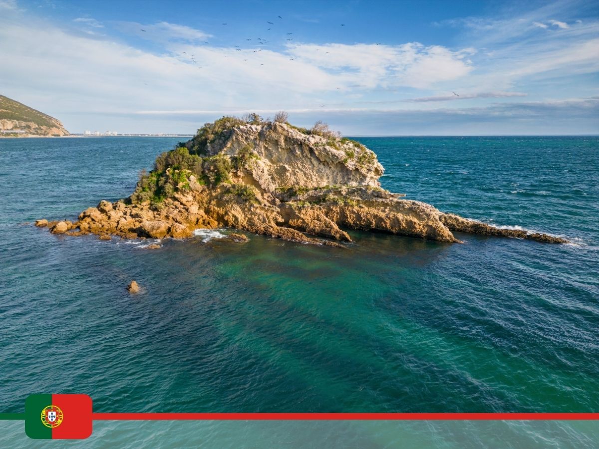 Small rocky island surrounded by blue-green ocean waters under a partly cloudy sky. There is a Portugal flag and a red line at the bottom of the image.