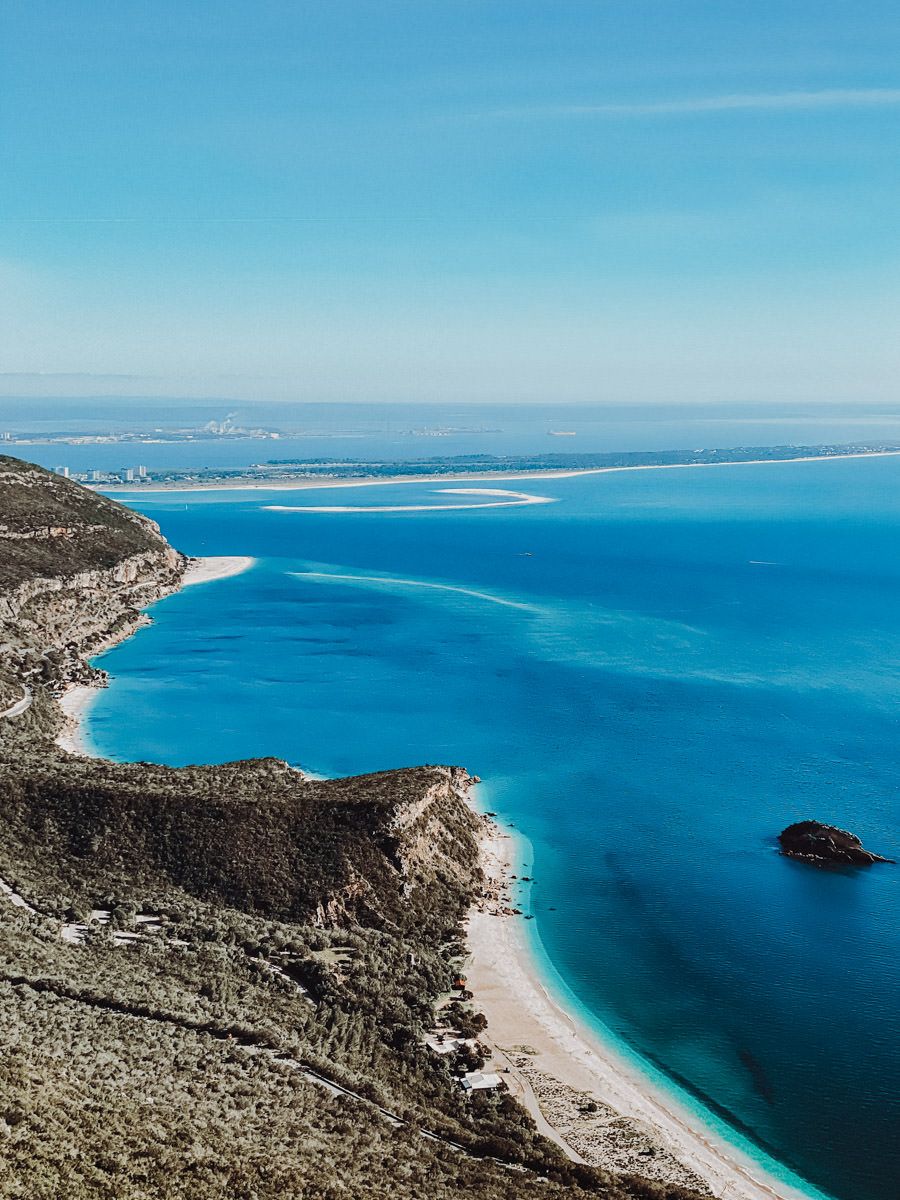 Aerial view of a coastline with clear blue water, sandy beaches, and rocky cliffs extending into the sea under a clear sky.