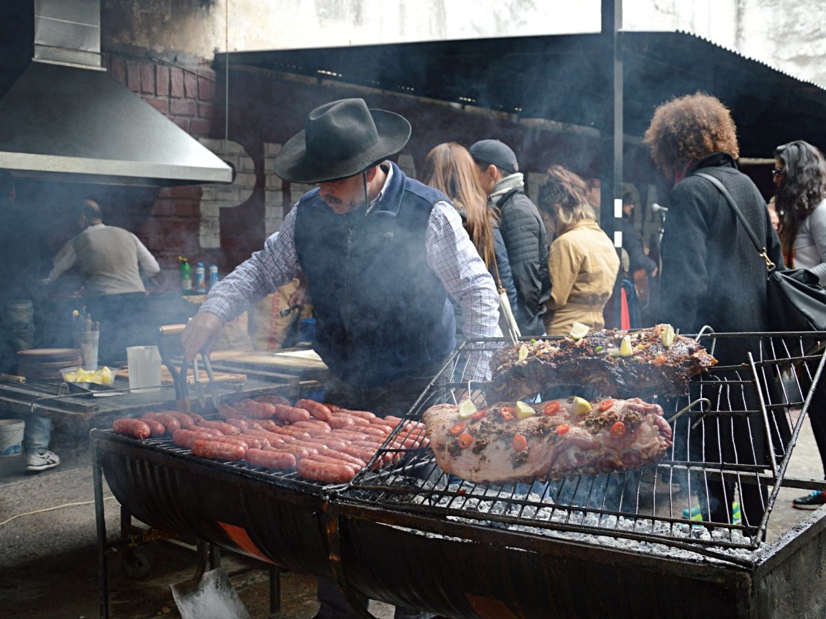 A man wearing a hat and vest grills sausages and large cuts of meat at an outdoor barbecue, surrounded by people.