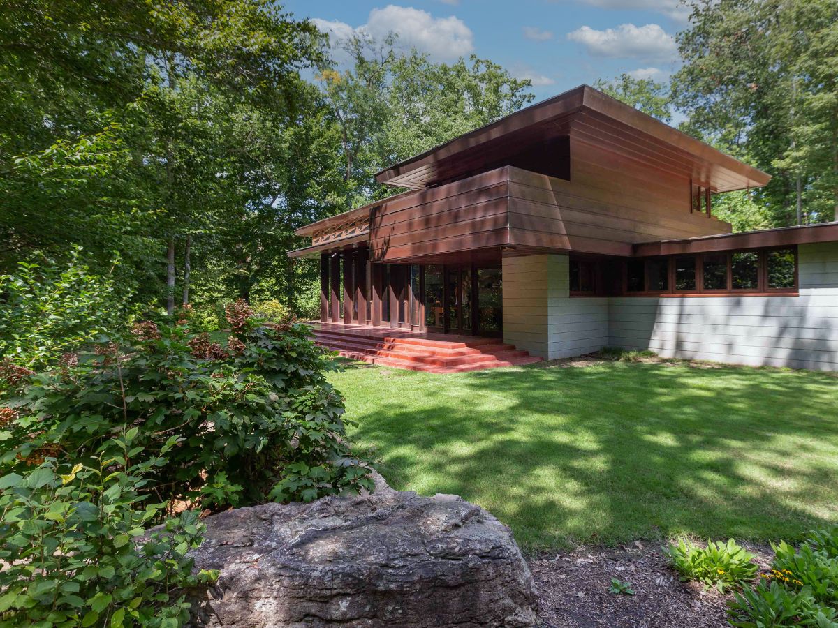 A modern house with large windows and a wooded backdrop, featuring a distinctive overhanging roof and a series of steps leading to the entrance, surrounded by well-maintained green lawns and foliage.