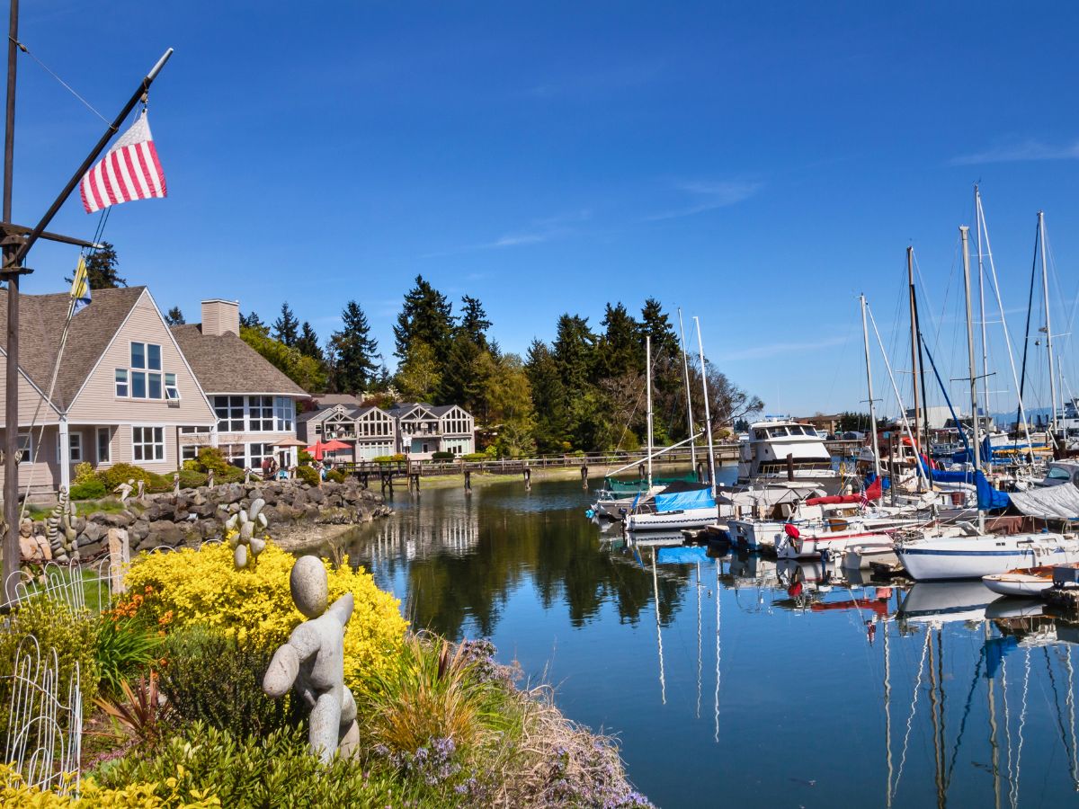 Marina scene with boats docked, waterfront houses, and an American flag. There's a garden with stone sculptures in the foreground, and a line of trees in the background under a clear blue sky.