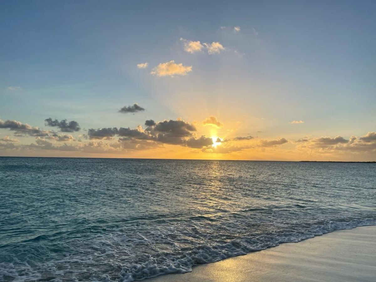 A serene beach at sunset with calm waves lapping onto the shore, and the sun setting behind partially cloudy skies.