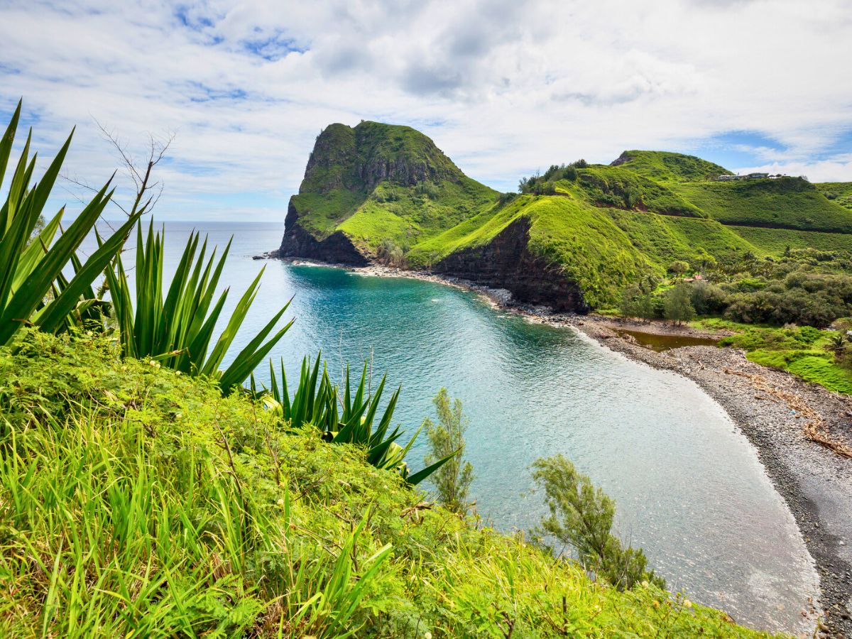 Scenic view of a lush green cliff beside a Maui beach under a cloudy sky.