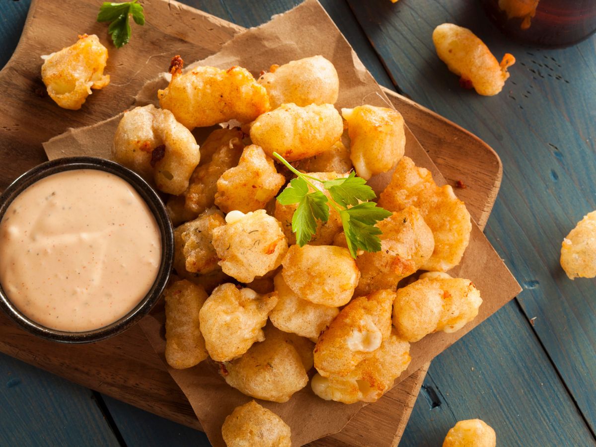 A pile of fried cheese curds on parchment paper, garnished with parsley, next to a small bowl of dipping sauce on a wooden surface.