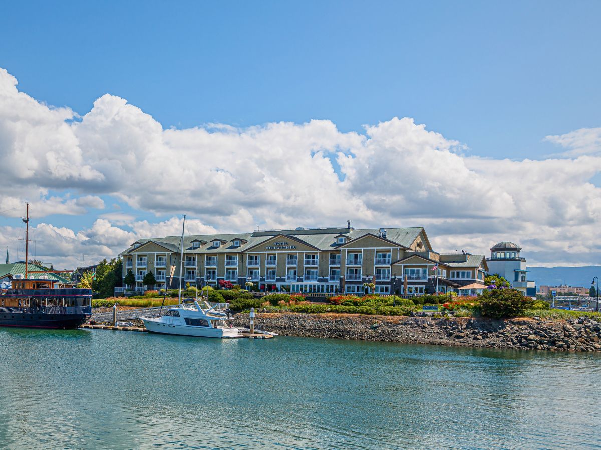 Waterfront hotel with a green roof by a marina, featuring a sailboat and motorboat docked in front. Blue sky with scattered clouds.