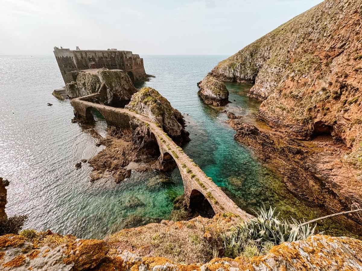 View of an old coastal fort connected by a stone bridge to a rocky cliff, surrounded by clear blue-green water. The fort and bridge are partially eroded, indicating age and exposure to elements.