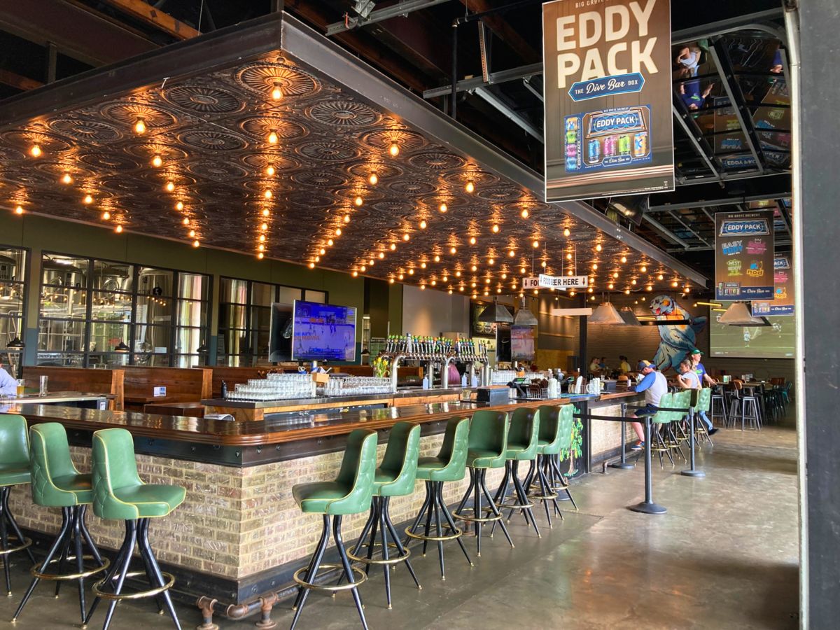 A restaurant and bar with a long counter, green bar stools, and hanging lights. People are seated at tables and a television is mounted on the wall.