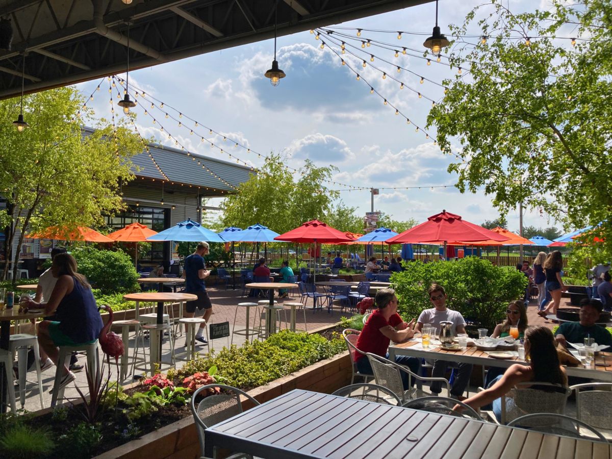 Outdoor restaurant patio with people dining, colorful umbrellas, and string lights hanging overhead. Trees and plants decorate the area with a building in the background on a sunny day.