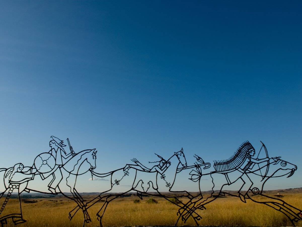 Metallic sculptures of native american hunters on horseback against a clear blue sky.