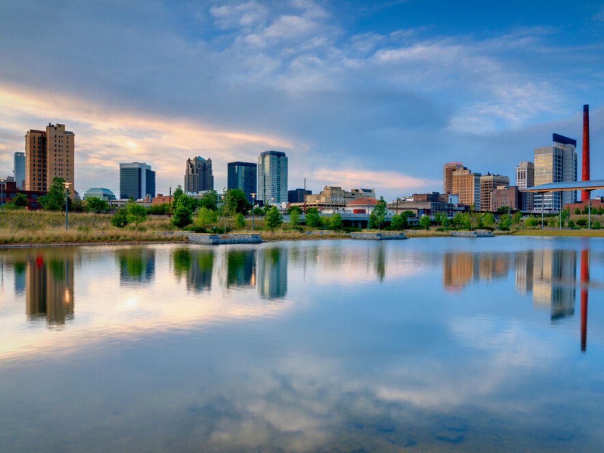 A city skyline featuring tall buildings reflected in a calm body of water under a blue sky with clouds.