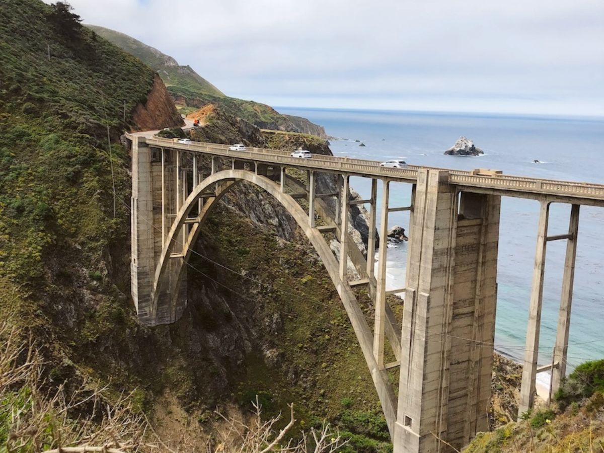 A concrete arch bridge spans a steep, green cliffside above the ocean with cars crossing, and the coastline is visible in the background.