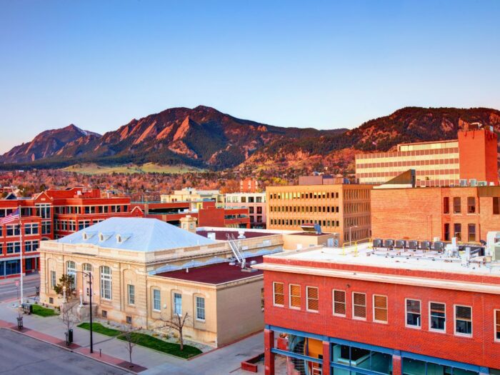 A cityscape view of Boulder, Colorado, featuring red-brick buildings with the Flatirons mountain range in the background under a clear sky.