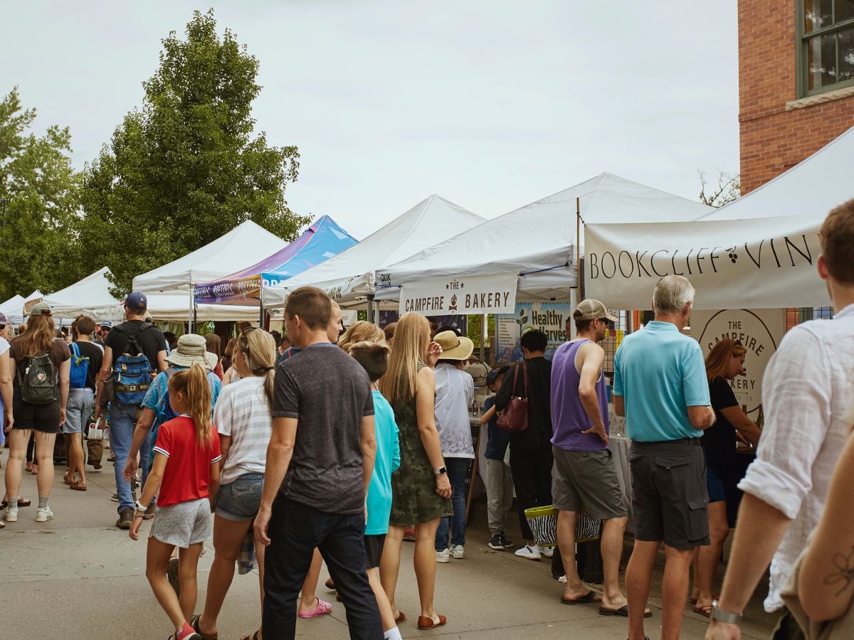 People browse booths at an outdoor market with various tents offering food and wine products. Trees and a brick building are visible in the background.