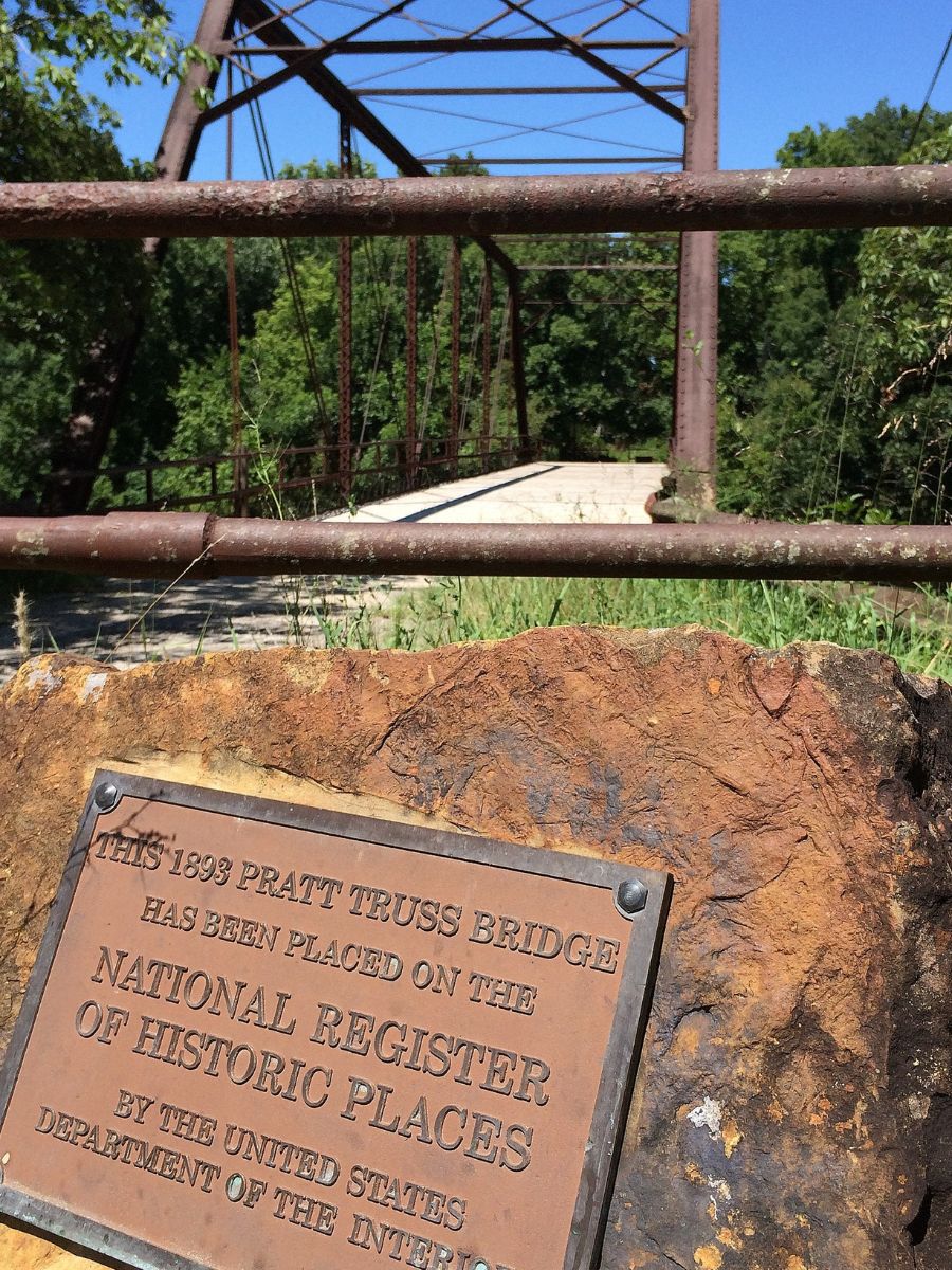 Plaque marks the 1898 Pratt Truss Bridge, listed on the National Register of Historic Places. Bridge and greenery visible in the background.