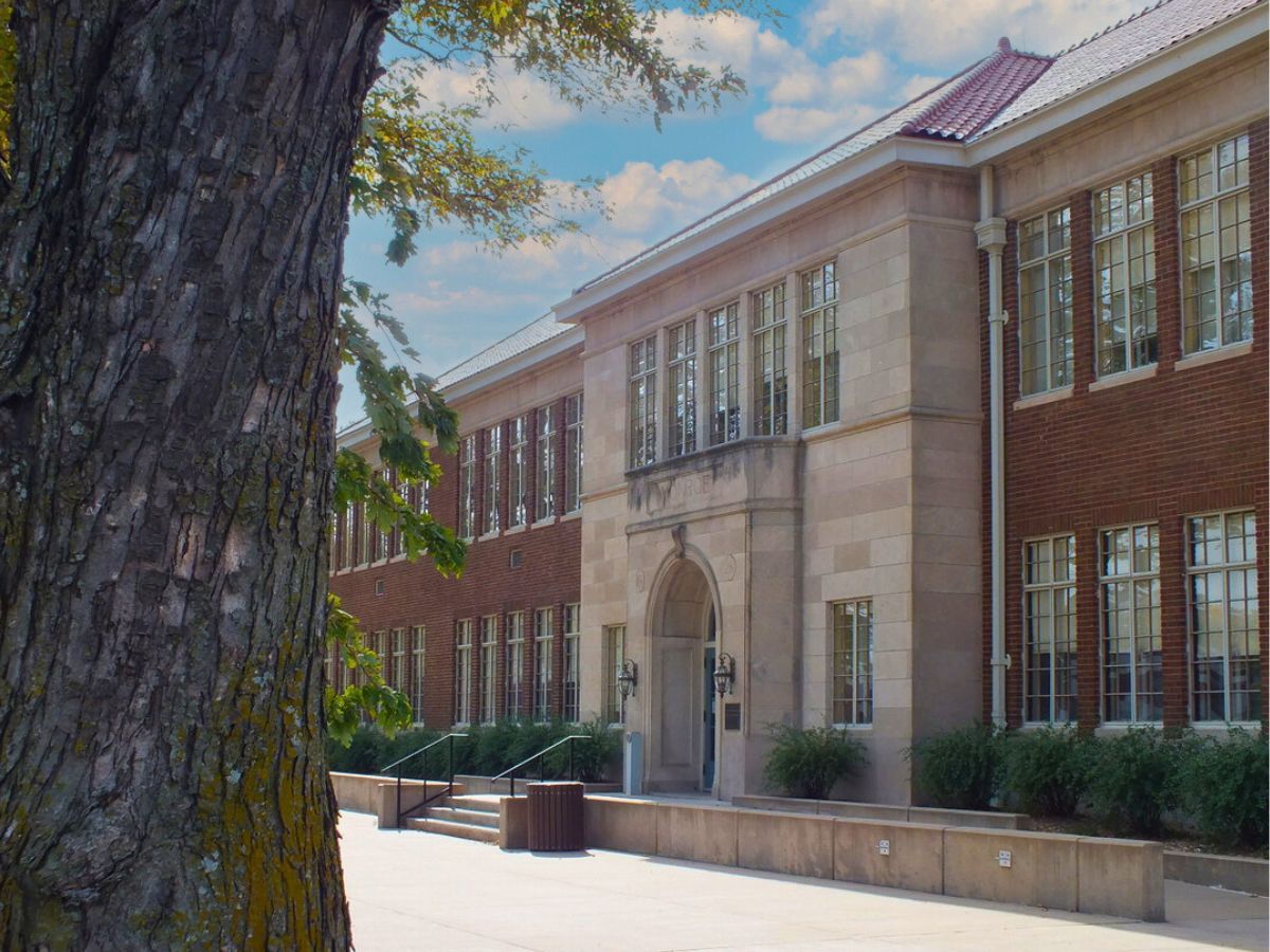 A large brick and stone building with multiple windows, a central arched entrance, and a tree in the foreground with clear skies in the background.