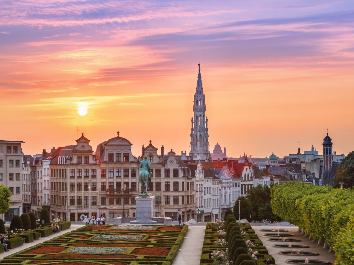 A view of a cityscape at sunset featuring a statue, intricately designed gardens, and a tall spire, with vibrant pink and orange hues in the sky.