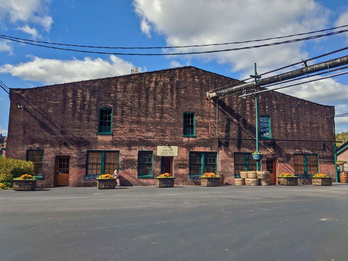 A large, old brick building with green-trimmed windows and doors, a sign in the center, metal pipes overhead, and planters with flowers in front, under a partly cloudy sky.