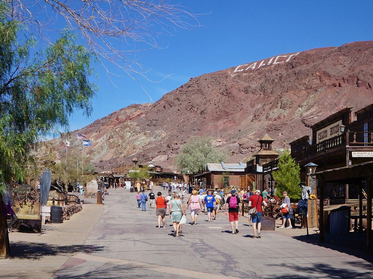 Visitors walk down a street in a desert town with brown hills and the word "Calico" written on a hillside.