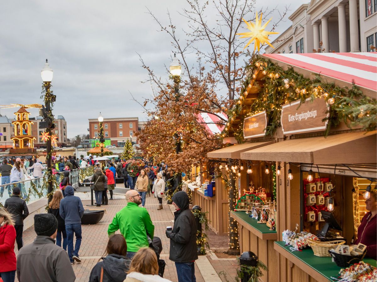 A busy outdoor holiday market with decorated wooden stalls, people walking, and festive lights on display. Trees and buildings are in the background.