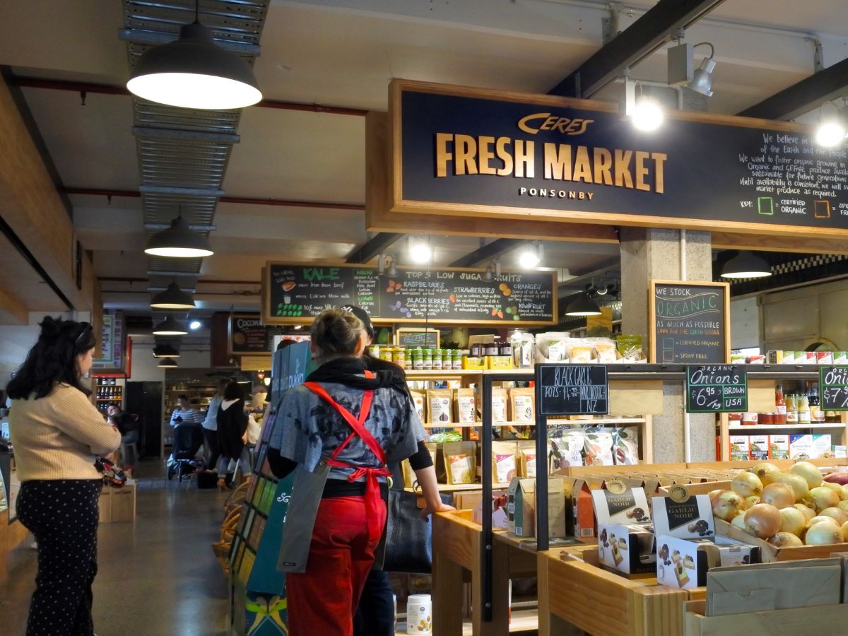 Shoppers browse in a well-lit, indoor market with various groceries and organic products displayed. A sign overhead reads "Ceres Fresh Market Ponsonby." Two women are visible near shelves of items.