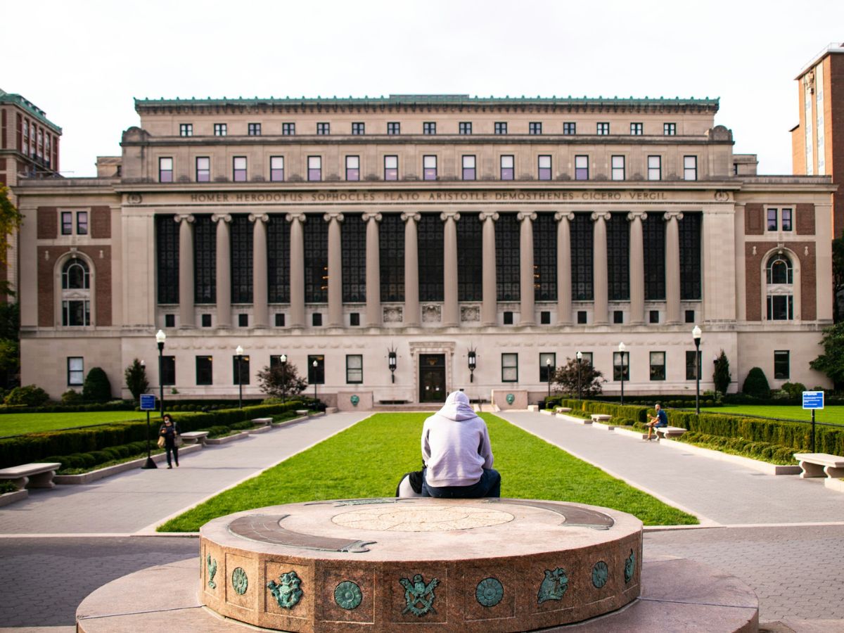 A person in a white hoodie sits on a circular stone structure facing a large university building with columns and inscriptions. The scene includes a green lawn and other people in the background.