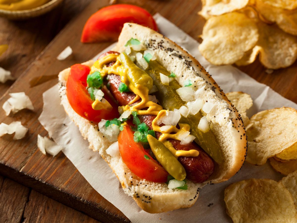 Chicago-style hot dog with mustard, onions, relish, tomato wedges, pickle spear, and sport peppers in a poppy seed bun, served alongside potato chips on a wooden board.