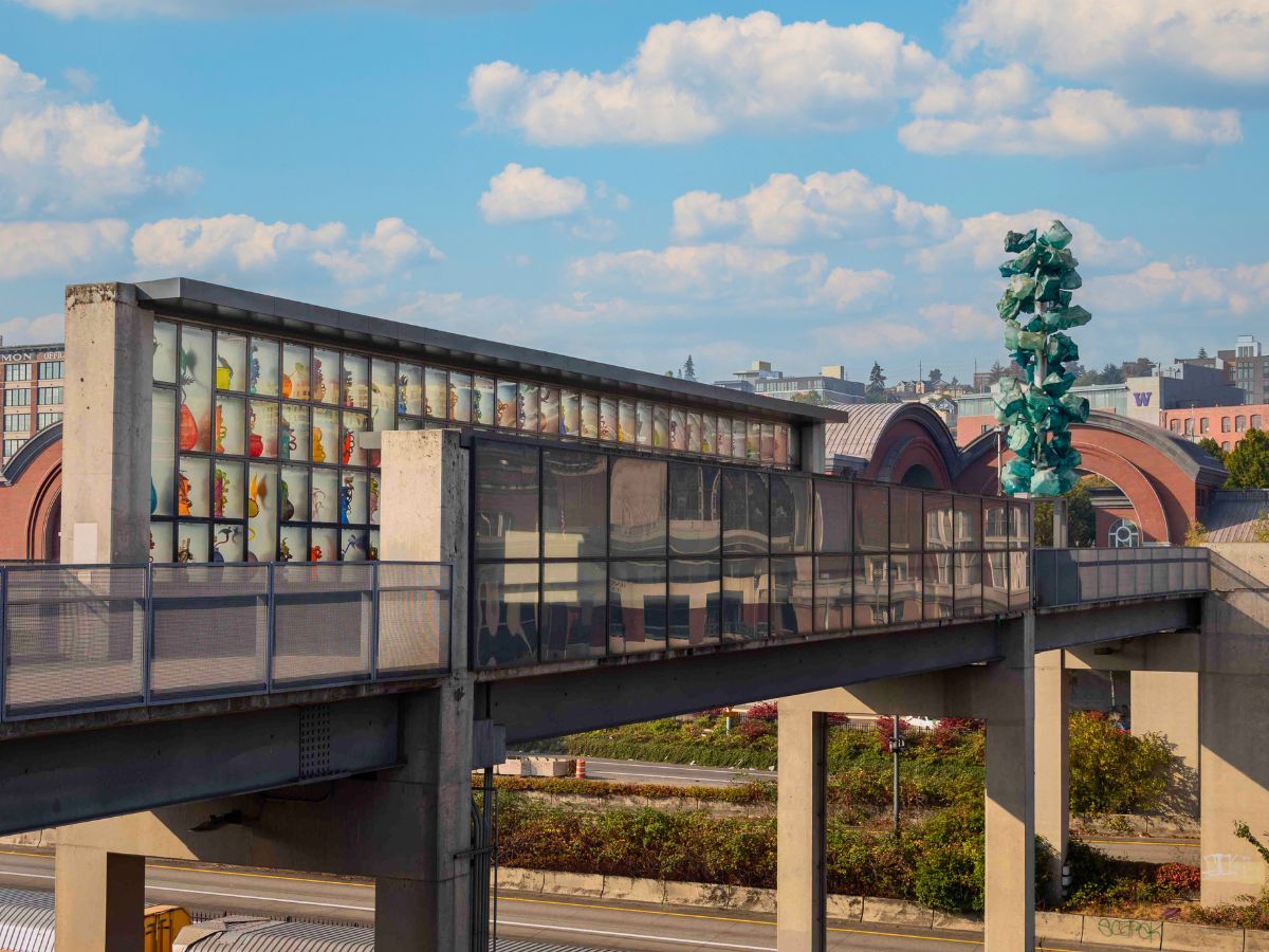 Pedestrian bridge with glass panels over a freeway, featuring colorful artwork and a tall green sculpture in the background.