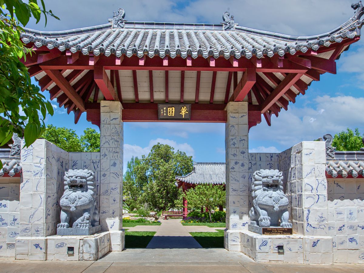 Traditional Chinese-style gate with two stone guardian lions at the entrance, surrounded by white-tiled walls featuring blue designs. Green trees and a courtyard are visible in the background.