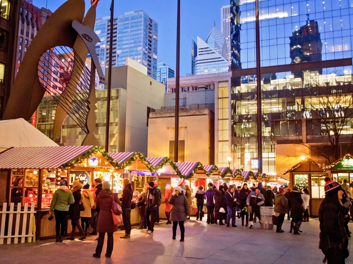 People browse vendors at an outdoor holiday market with decorated stalls, surrounded by city skyscrapers and a large sculpture.