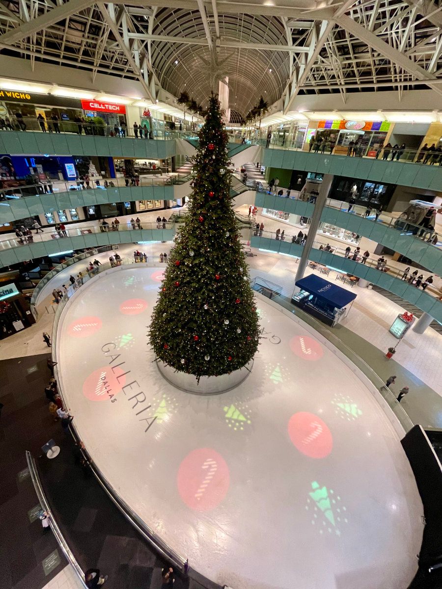 A tall Christmas tree stands at the center of an indoor ice skating rink within a multi-level shopping mall.