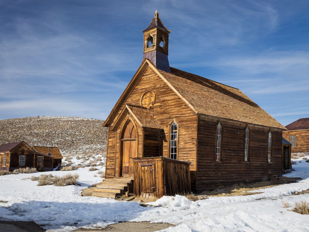 A weathered wooden church stands in a snowy landscape with clear skies. Nearby are a few wooden structures on a slightly hilly terrain.
