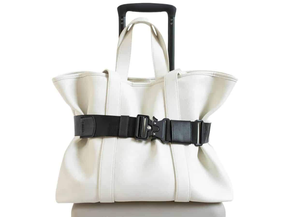 A black Cincha travel belt holding a white shoulder bag onto a roll-on suitcase.