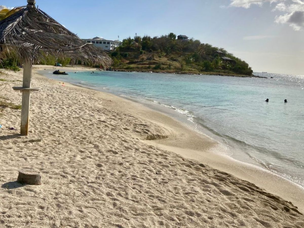 A sandy beach with a thatched umbrella, clear blue water, and a small hill in the background. A few people are swimming.