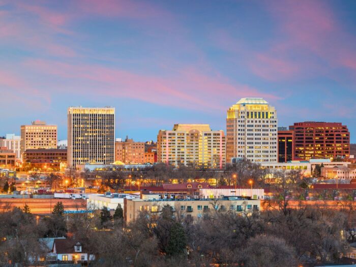 Skyline of a city at dusk with lit buildings against a backdrop of a pink and blue sky. Trees and lower buildings are visible in the foreground.