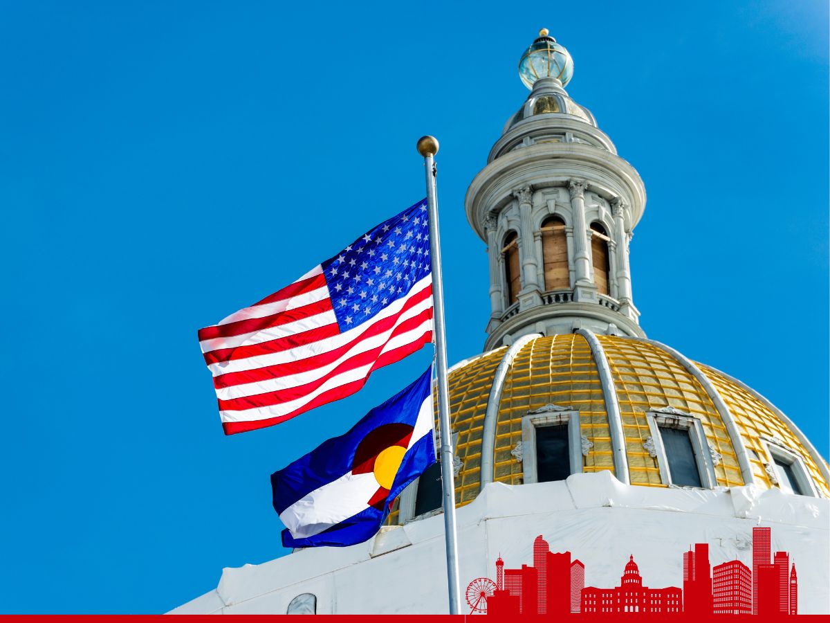 View of the United States and Colorado flags waving in front of the dome of the Colorado State Capitol under a clear blue sky.