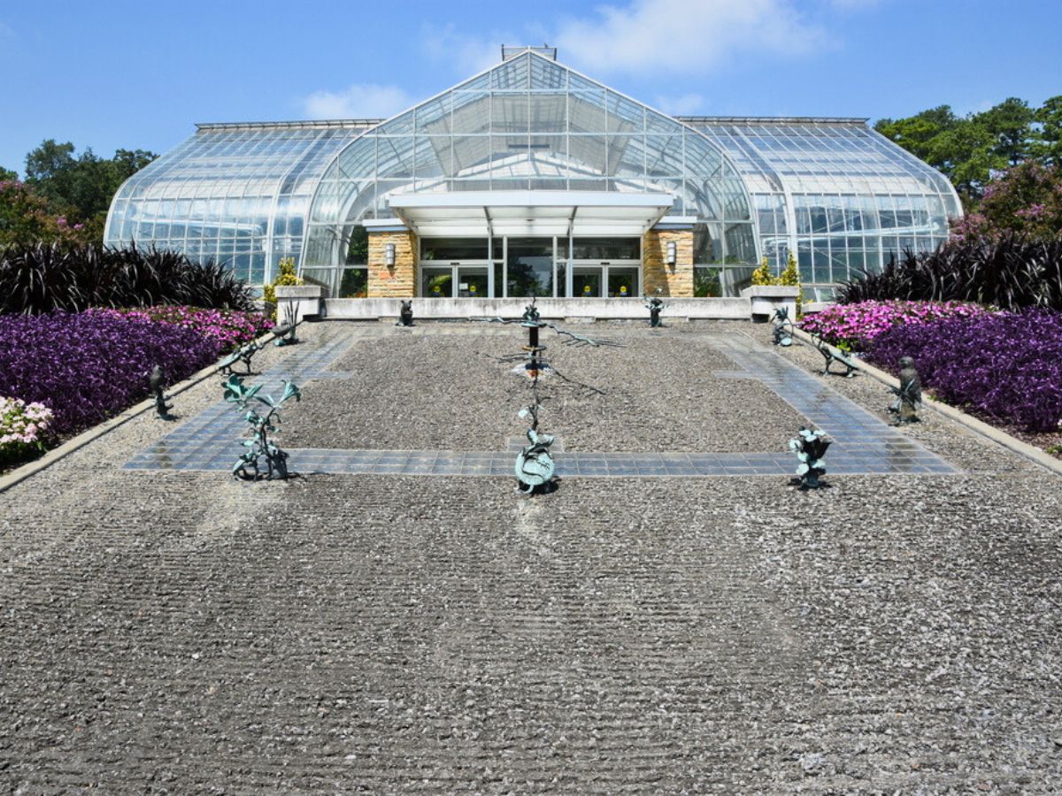 The conservatory at the Birmingham Botanical Gardens is a large greenhouse with a glass facade and metal framework, surrounded by vibrant purple and green landscaping.