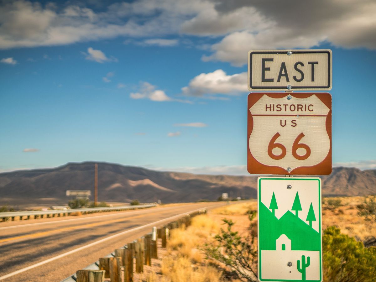 Road sign for Historic Route 66 heading east, with a scenic desert landscape and mountains in the background.