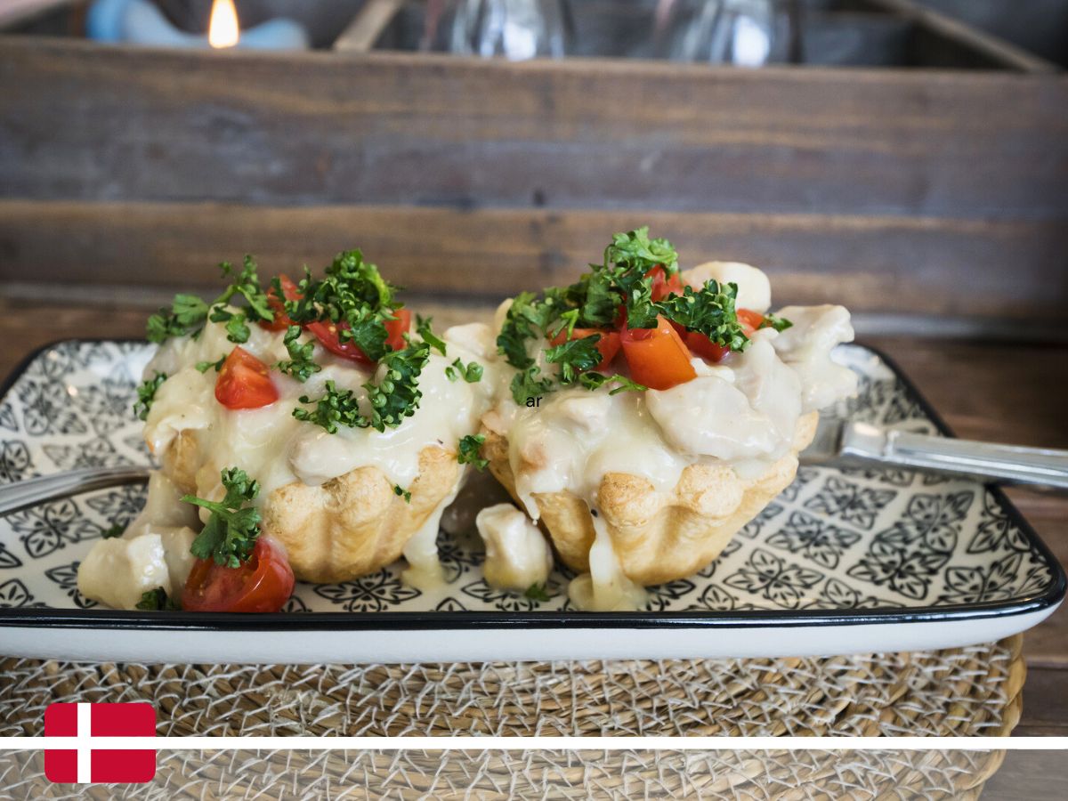 Two small pastry tarts filled with a creamy white sauce and topped with chopped tomatoes and parsley, served on a patterned rectangular plate with one fork on the side. Danish flag in bottom left corner.