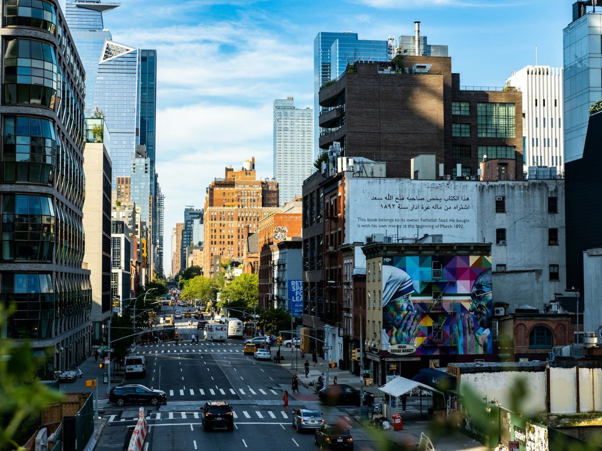 City street lined with buildings, including a mural and a billboard with Arabic text. Cars and pedestrians move along the street under a clear blue sky.