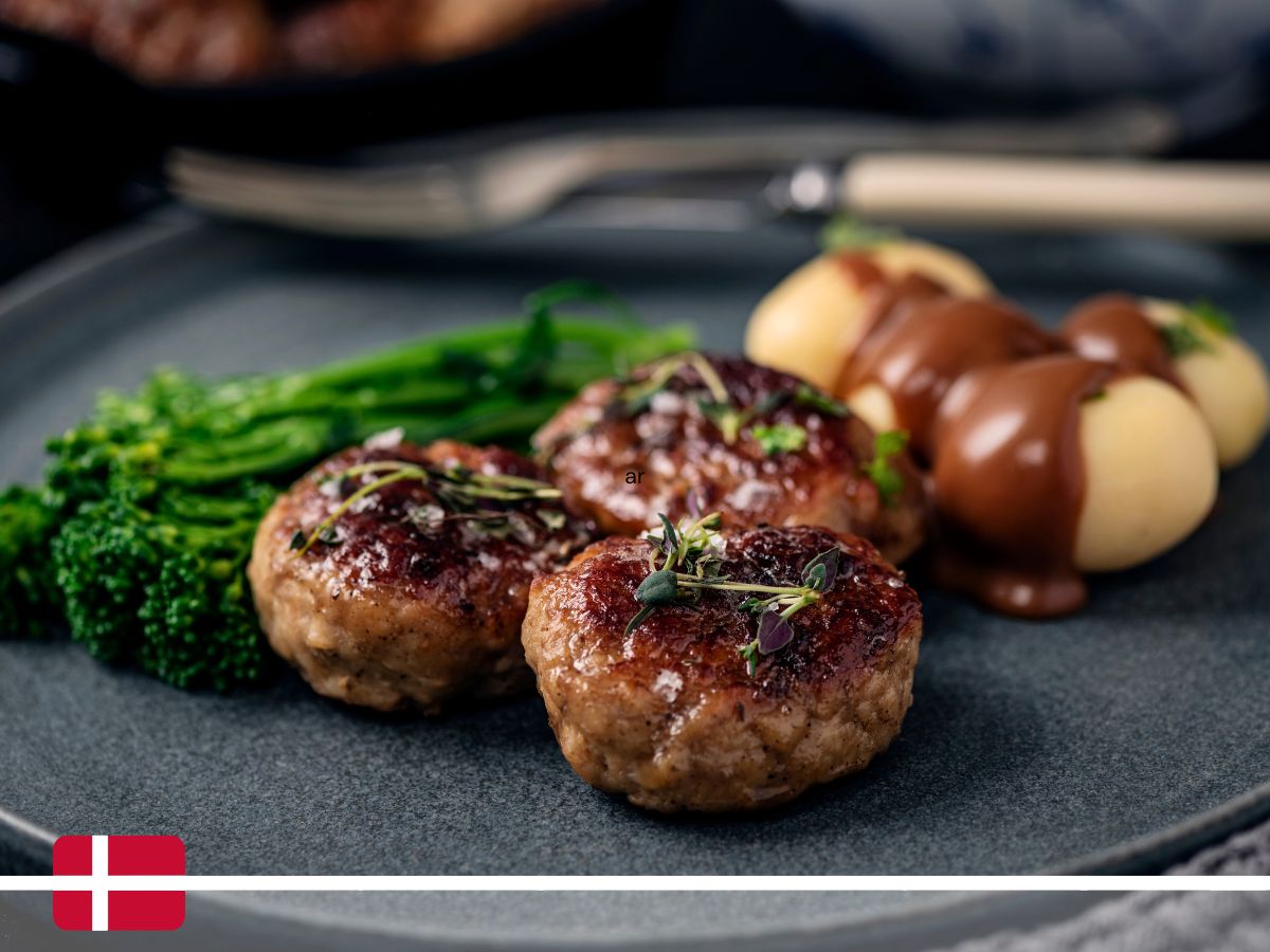 A plate with three meatballs garnished with herbs, steamed broccoli, and boiled potatoes topped with brown gravy. A fork and knife are placed behind the plate. Danish flag icon in the lower-left corner.