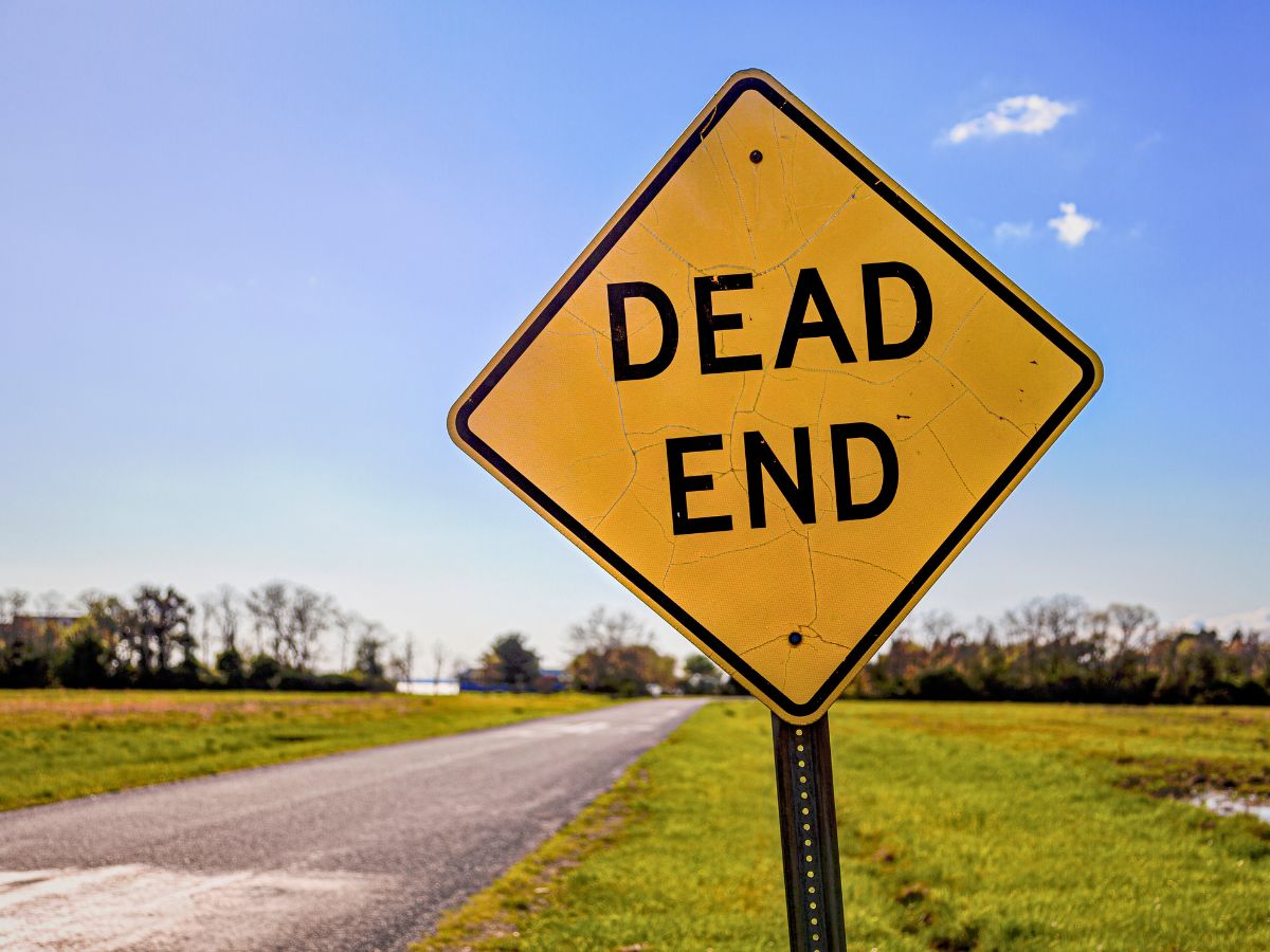 Yellow "Dead End" sign on a rural road with greenery and blue sky in the background.